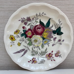 Könnte beinhalten: Weißer Keramikteller mit gewelltem Rand, verziert mit einem Blumenstrauß. Der Strauß zeigt rote, gelbe, lila und weiße Blumen mit grünen Blättern. Ein dekoratives Stück zum Servieren.