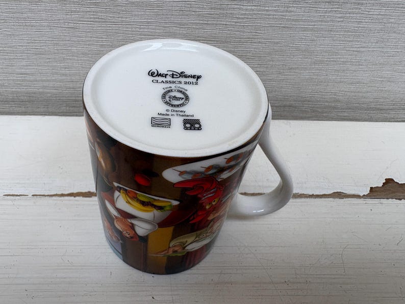 May include: White ceramic mug featuring a colorful illustration of characters from a Disney movie. The mug has a curved handle and the text "Walt Disney Classics 2012" is printed on the bottom.