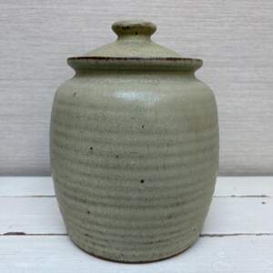 May include: A handmade, light green ceramic jar with a lid. The jar has a rounded body with horizontal ridges and a small, rounded lid. The jar is approximately 6 inches tall and 4 inches wide.