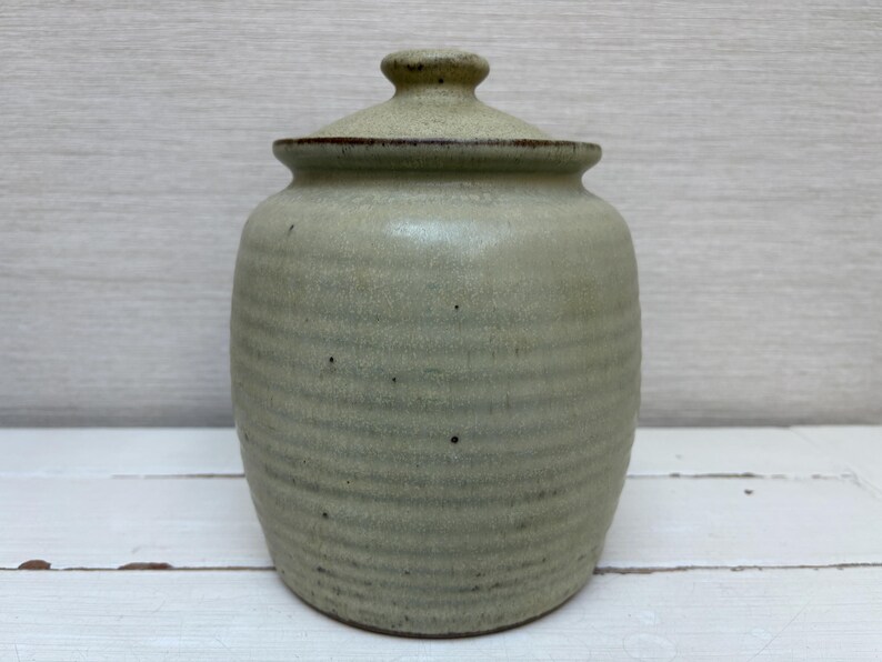 May include: A handmade, light green ceramic jar with a lid. The jar has a rounded body with horizontal ridges and a small, rounded lid. The jar is approximately 6 inches tall and 4 inches wide.