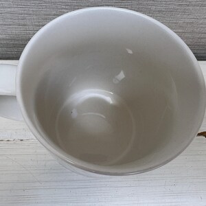 May include: A white ceramic mug with a curved handle. The mug is empty and has a glossy finish. The interior of the mug is visible, showing a smooth, rounded surface. The mug is sitting on a white wooden surface.