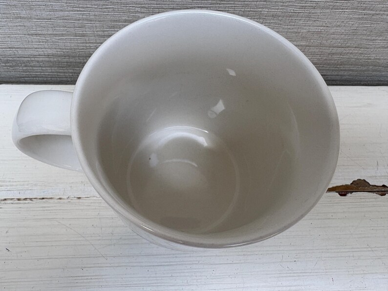 May include: A white ceramic mug with a curved handle. The mug is empty and has a glossy finish. The interior of the mug is visible, showing a smooth, rounded surface. The mug is sitting on a white wooden surface.