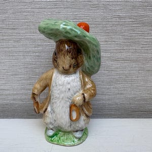 Può includere: Statuina in ceramica di un coniglio con un cappello verde e una piccola borsa. Il coniglio ha la pelliccia marrone, il petto bianco e si trova su un pezzo d'erba verde. Il cappello ha un piccolo dettaglio arancione.