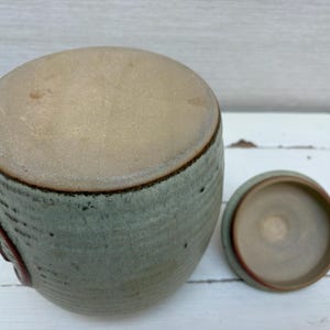 May include: A ceramic jar with a lid, in shades of green and tan. The jar has a rounded shape with a textured surface and a dark brown rim. The lid is off to the side, revealing the interior. The jar is on a white surface.