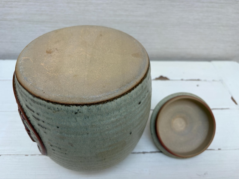 May include: A ceramic jar with a lid, in shades of green and tan. The jar has a rounded shape with a textured surface and a dark brown rim. The lid is off to the side, revealing the interior. The jar is on a white surface.