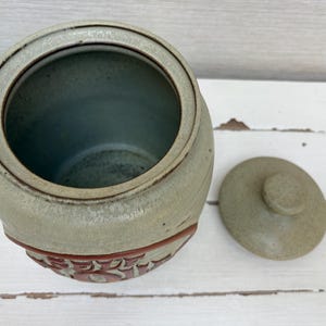 May include: A ceramic jar with a lid, in shades of green and brown. The jar has a rounded shape with a decorative band of red floral designs. The lid is round with a small knob. The jar is empty.