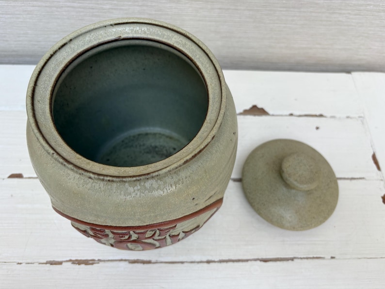May include: A ceramic jar with a lid, in shades of green and brown. The jar has a rounded shape with a decorative band of red floral designs. The lid is round with a small knob. The jar is empty.