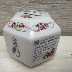 Op de afbeelding: Witte keramische spaarpot met een zeshoekige vorm, versierd met illustraties van Peter Rabbit en bloemmotieven. De tekst "For your christening" is zichtbaar aan de bovenkant. Decoratieve linten en tekst uit een verhaal.