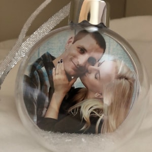May include: A clear glass Christmas ornament with a silver top and a ribbon. Inside the ornament is a photo of a couple kissing. The ornament is filled with white glitter.