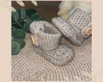 May include: A pair of handmade, light gray crochet baby booties. Each bootie features a folded cuff and a wooden button closure. The booties are displayed on a textured, neutral-toned surface, suggesting a cozy, handcrafted theme.