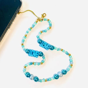 Puede incluir: Un collar de cuentas azules y verdes con detalles dorados y dos abalorios azules con la palabra "LOVE". El collar está sobre un fondo blanco.