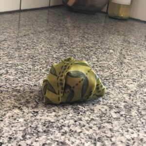 May include: A folded green fabric wrap with a leaf pattern. The wrap is a light green color with dark green leaf and dot designs. The fabric is folded into a small bundle.