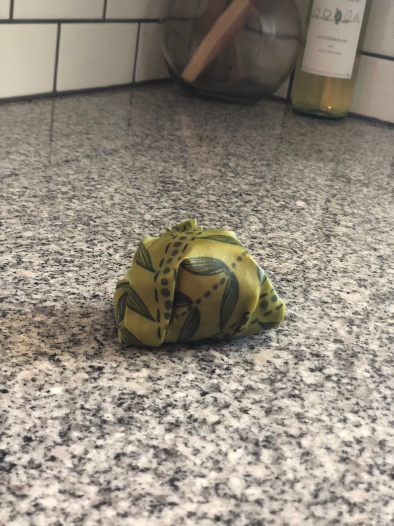 May include: A folded green fabric wrap with a leaf pattern. The wrap is a light green color with dark green leaf and dot designs. The fabric is folded into a small bundle.