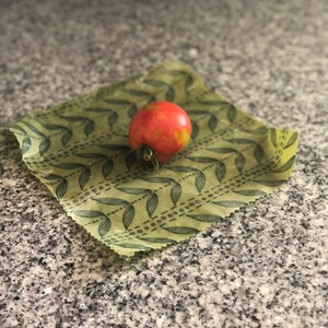 May include: A ripe red tomato with green stem rests on a reusable beeswax food wrap. The wrap is light green with a repeating pattern of dark green leaves. The background is a speckled gray surface.