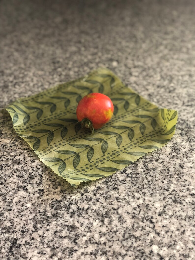 May include: A ripe red tomato with green stem rests on a reusable beeswax food wrap. The wrap is light green with a repeating pattern of dark green leaves. The background is a speckled gray surface.