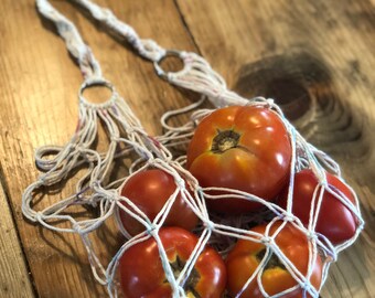 May include: A white macrame net bag filled with ripe red tomatoes. The bag has a long handle and metal rings. The tomatoes are various sizes and shapes, with green stems. The bag is sitting on a wooden surface.