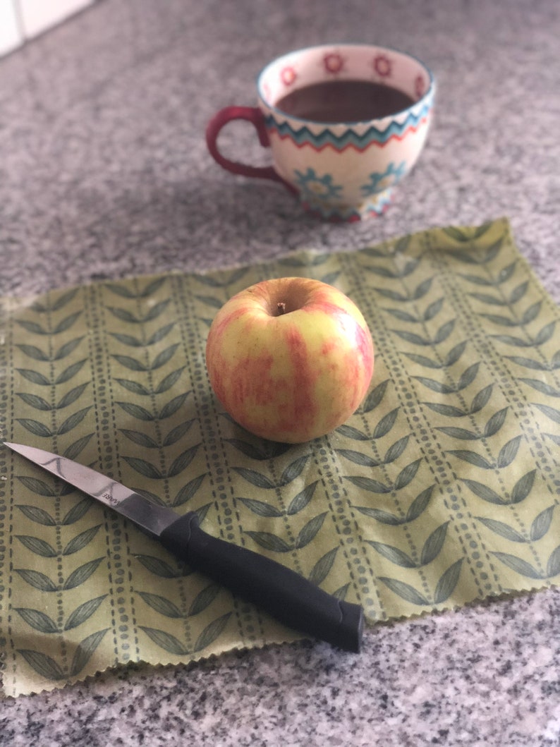 May include: A red and yellow apple rests on a green leaf-patterned wrap next to a small knife with a black handle. A floral-decorated mug filled with a dark liquid sits in the background. The scene is set on a speckled countertop.