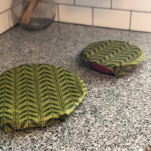 May include: Two reusable food wraps with a green leaf pattern. One wrap covers a red bowl, and the other is placed on the counter. The wraps are made of a flexible material and are designed to replace plastic wrap.