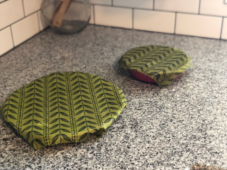 May include: Two reusable food wraps with a green leaf pattern. One wrap covers a red bowl, and the other is placed on the counter. The wraps are made of a flexible material and are designed to replace plastic wrap.