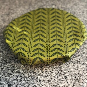 May include: A round, green fabric bowl cover with a repeating pattern of black leaves and dotted lines. The cover is made of a soft, flexible material and is designed to fit over bowls or containers. The cover is on a gray granite countertop.