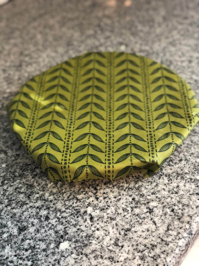 May include: A round, green fabric bowl cover with a repeating pattern of black leaves and dotted lines. The cover is made of a soft, flexible material and is designed to fit over bowls or containers. The cover is on a gray granite countertop.