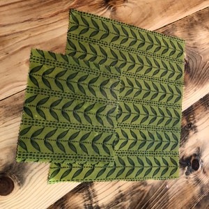May include: Two green beeswax food wraps with a black leaf and dot pattern. The wraps are rectangular and vary slightly in size. They are placed on a wooden surface with a natural wood grain.