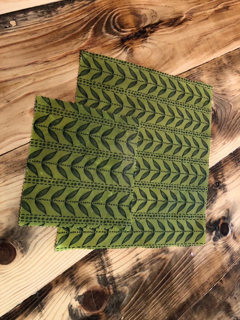 May include: Two green beeswax food wraps with a black leaf and dot pattern. The wraps are rectangular and vary slightly in size. They are placed on a wooden surface with a natural wood grain.