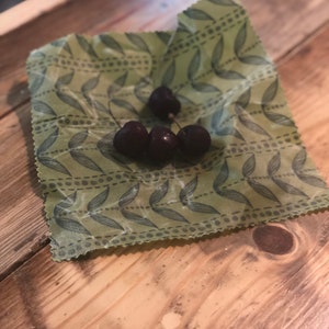 May include: A close-up of four dark red cherries on a green beeswax food wrap with a leaf pattern. The wrap is on a wooden surface. The cherries are fresh and ripe.
