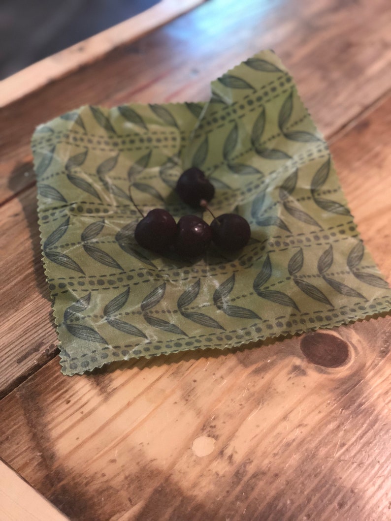 May include: A close-up of four dark red cherries on a green beeswax food wrap with a leaf pattern. The wrap is on a wooden surface. The cherries are fresh and ripe.