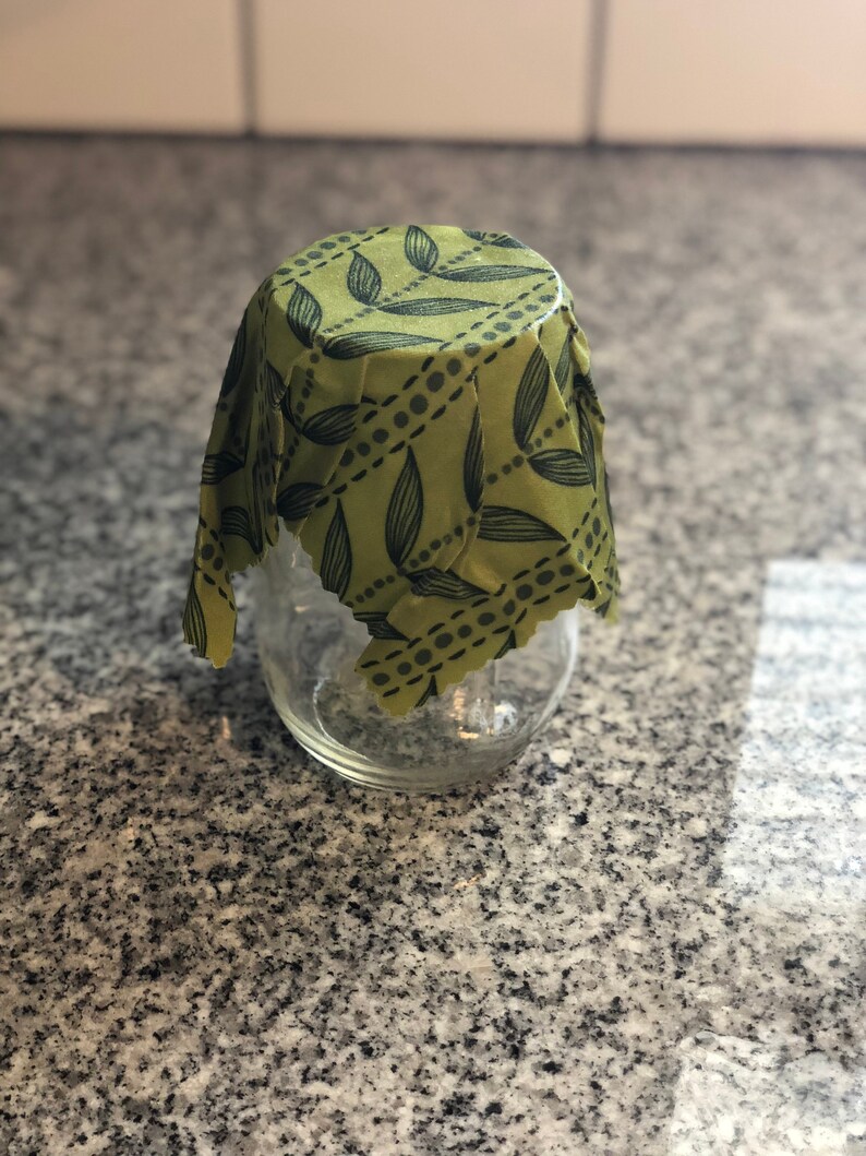 May include: A clear glass jar covered with a green beeswax food wrap. The wrap has a pattern of black leaves and dotted lines. The jar sits on a speckled gray countertop.