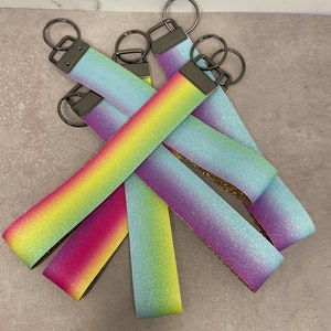 May include: Five rainbow ombre glitter keychains with silver key rings. The keychains are made of fabric and have a gradient of colors, including pink, yellow, green, blue, and purple.