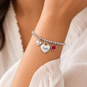 Puede incluir: Una pulsera de cuentas plateadas con tres dijes: un corazón pequeño grabado con el número 30, un corazón más grande grabado con el nombre Sophia y un dije circular con una piedra preciosa redonda, facetada y de color rojo vibrante.