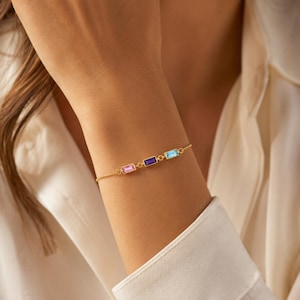 May include: A delicate gold chain bracelet featuring three rectangular baguette-cut gemstones in a row. The stones are set in gold bezels and include a light pink, a deep purple, and a bright blue gemstone, connected by thin gold links.