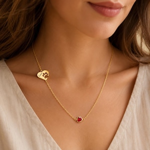 Può includere: Una collana a catena color oro con un ciondolo a forma di cuore con un'impronta di zampa ritagliata e il nome Luna inciso, abbinata a un secondo ciondolo più piccolo a forma di cuore con una singola pietra preziosa rossa incastonata.