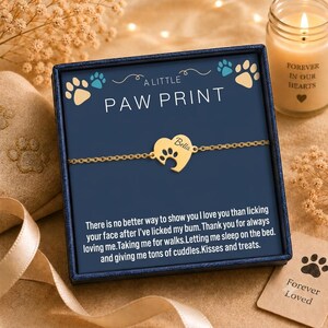 May include: A gold-toned chain bracelet featuring a heart-shaped charm with a paw print cutout and the name Bella engraved on it. The bracelet is presented in a dark blue gift box with the text A LITTLE PAW PRINT and a printed message.