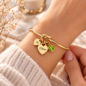 May include: A gold-toned bangle bracelet featuring a knot detail and three dangling charms: a small heart engraved with the number 30, a larger heart engraved with the name Olivia, and a round, faceted lime green gemstone charm.