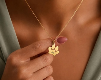May include: A gold-toned necklace featuring a delicate chain and a pendant shaped like a paw print. The center of the paw print pendant is engraved with the name Bella in a cursive script font. The metal has a smooth, polished finish.