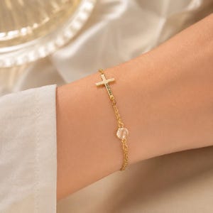 Puede incluir: Una pulsera de cadena en tono dorado que presenta un pequeño dije de cruz grabado con la fecha 10.03.2001 y una piedra redonda transparente. La delicada cadena de metal conecta la cruz y la piedra, creando una pieza de joyería sencilla.