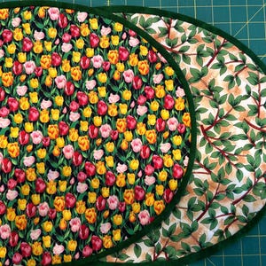 Könnte beinhalten: Zwei ovale Tischsets. Eines zeigt ein Muster aus roten, rosa und gelben Tulpen auf schwarzem Hintergrund mit einem grünen Rand. Das andere hat ein Muster aus grünen Blättern und braunen Zweigen auf cremefarbenem Hintergrund, ebenfalls mit einem grünen Rand.