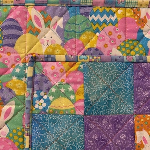 May include: A colorful Easter-themed quilt featuring bunnies and decorated eggs. The quilt has a patchwork design with various patterns and colors, including pink, purple, yellow, and blue. The edges are finished with a patterned border.