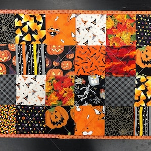 Könnte beinhalten: Eine rechteckige, gesteppte Stoffmatte mit einem Patchwork-Design aus verschiedenen Halloween-Motiven, darunter Kürbisse, Zuckermais, Spinnennetze, Skelette, Herbstblätter und Hexenhüte, eingefasst mit einem orangefarbenen Stoffrand.