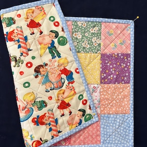 Könnte beinhalten: Zwei gesteppte Topflappen. Einer zeigt einen Vintage-Druck von Kindern mit Eis und Süßigkeiten auf cremefarbenem Hintergrund, umrandet von hellblauem Stoff. Der andere hat ein Patchwork-Design mit Blumen- und Karomustern in verschiedenen Farben, ebenfalls mit hellblauem Rand.