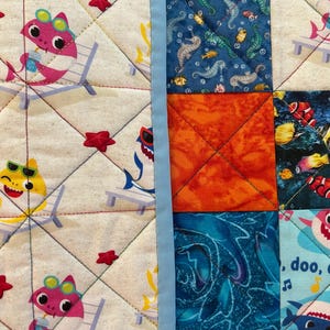 May include: A quilted fabric featuring a colorful pattern of cartoon sharks in sunglasses relaxing on beach chairs, interspersed with red star shapes. The design includes squares with seahorses, fish, and an orange square.
