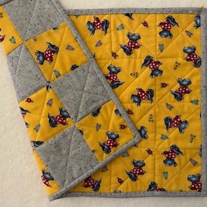 May include: A quilted placemat and coaster set. The placemat features a yellow background with a repeating pattern of Scottish Terrier dogs wearing red polka-dot sweaters. The border and the coaster are gray.