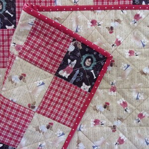 May include: A quilted patchwork featuring a dog theme. The design incorporates squares of red plaid, beige fabric with dog illustrations, and a black square with a dog wearing a sweater. The edges are finished with a red border.