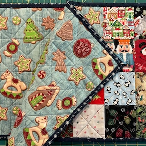 May include: A quilted Christmas placemat with a light blue background featuring gingerbread men, stars, and Christmas tree designs. The corner has a patchwork design with Santa, snowmen, and penguin patterns.