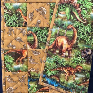 Könnte beinhalten: Eine braune und beige gesteppte Stofftischdecke mit Dinosaurier-Motiv. Die Tischdecke zeigt verschiedene Dinosaurier, darunter einen grünen Stegosaurus, einen braunen Brachiosaurus und einen blauen Velociraptor. Die Tischdecke ist aus einem weichen, bequemen Stoff gefertigt und perfekt, um jedem Esstisch eine lustige Note zu verleihen.