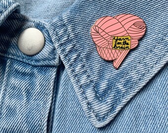 Op de afbeelding: Een roze emaille pin in de vorm van een brein gemaakt van garen, met de woorden "Yarn on the Brain" op een gele vierkant. De pin is geplaatst tegen een blauwe denim achtergrond.