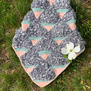 May include: A crocheted blanket with a geometric pattern in shades of gray, pink, and green. The blanket is made with a chunky yarn and has a soft, textured feel.