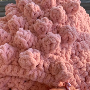 May include: Close-up of a pink crocheted blanket being made with a pink crochet hook. The blanket has a textured, puffy design.
